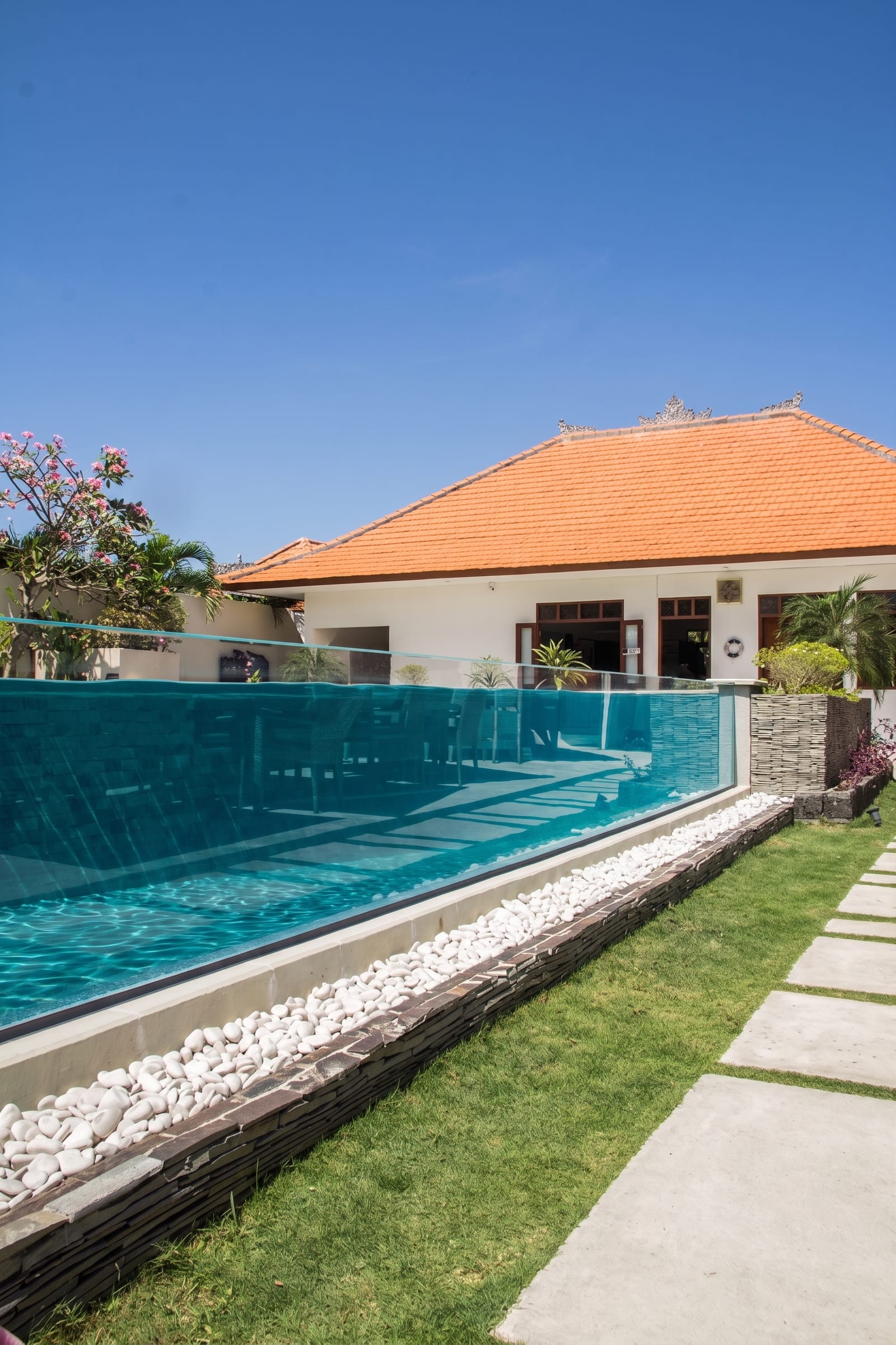 Glass-walled overflow pool with clear water and submerged steps, edged by a white-pebble and natural-stone border beside a lawn at a Balinese villa