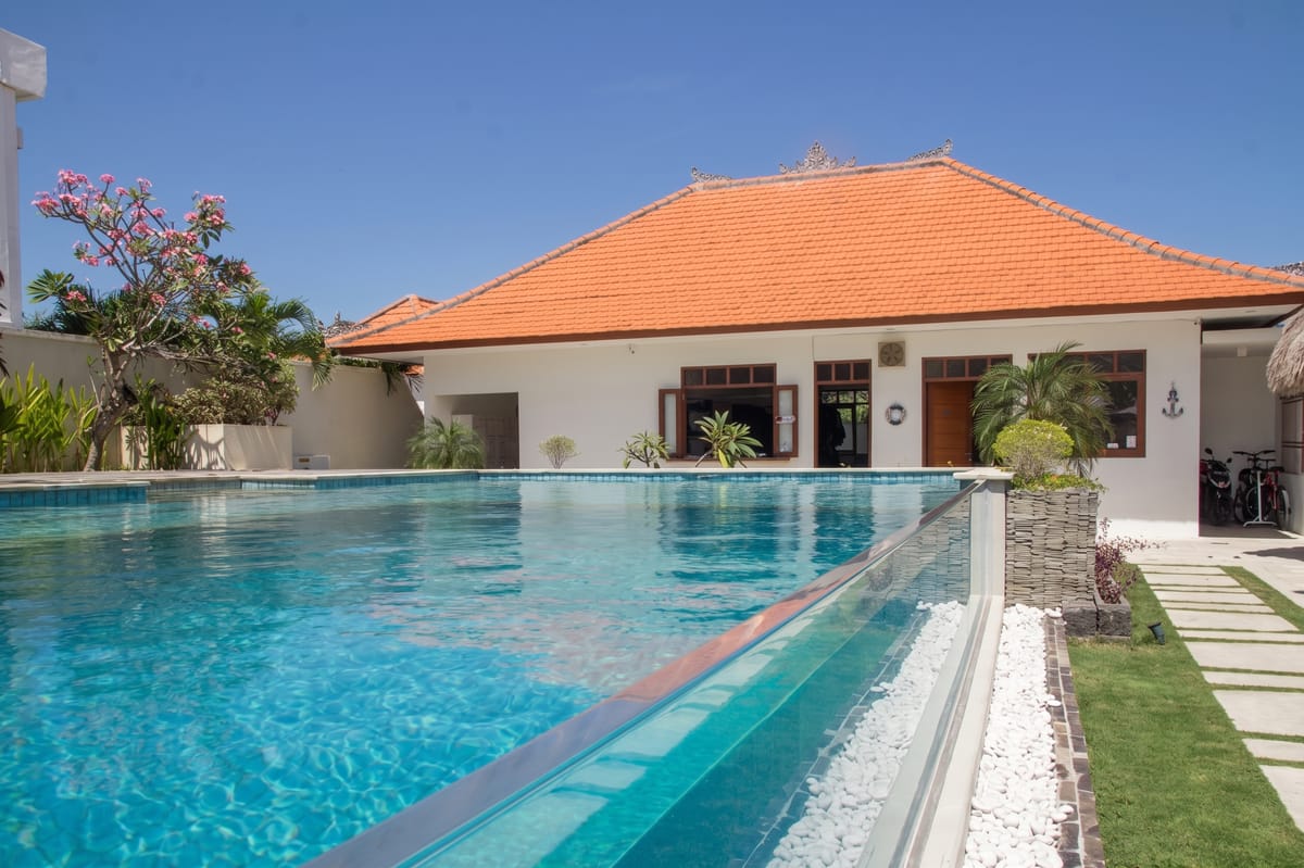 Wide view of a glass-walled overflow pool with a white-pebble and stone border and a stepping-stone lawn beside a Balinese villa with an orange tile roof