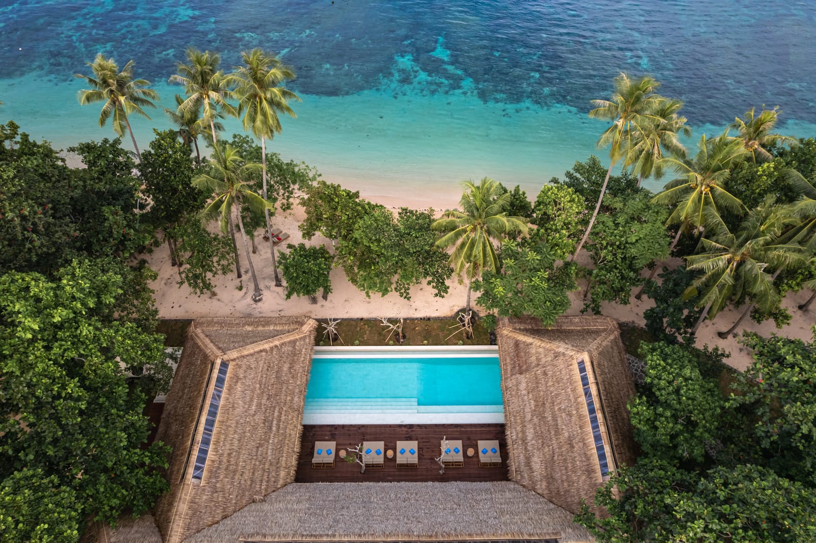 Aerial view of a beachfront pool set between thatched pavilions, fringed by palms above a white-sand beach and turquoise reef at a private island resort in Indonesia