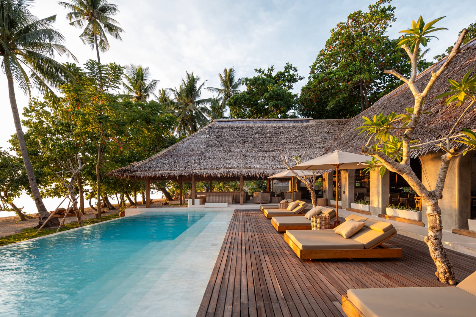 Resort pool alongside a timber deck with daybeds and a parasol beside a thatched beach bar and palms at golden hour on a private island resort in Indonesia