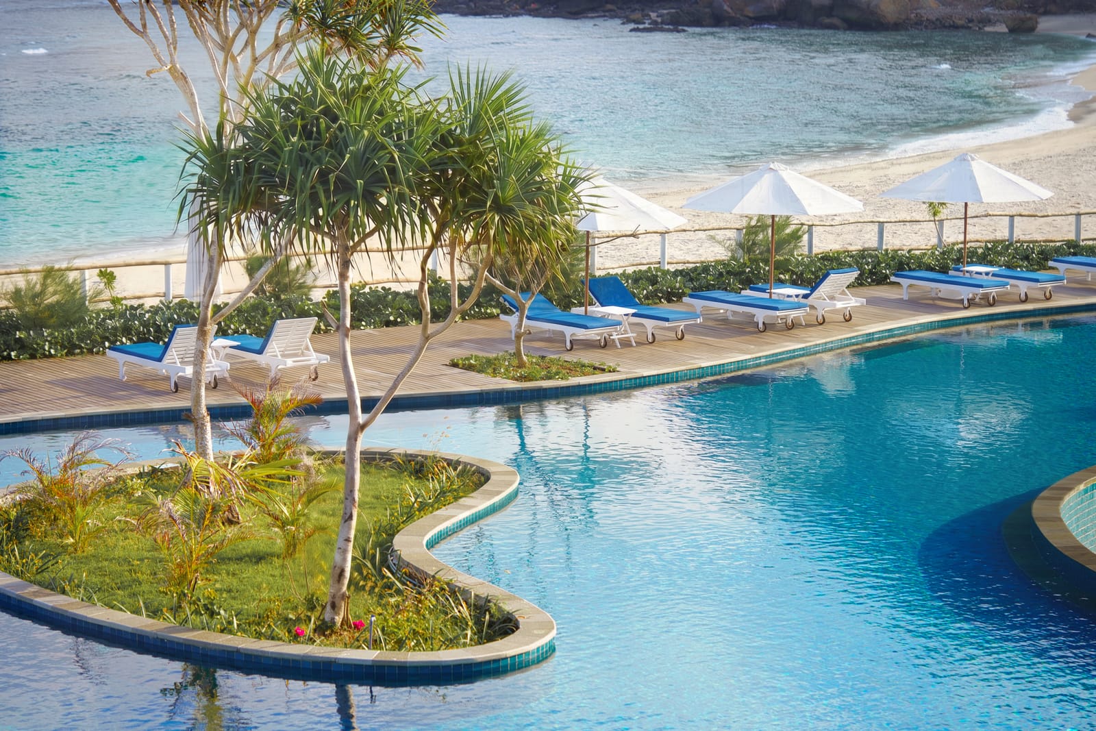 Beachfront lagoon pool with white parasols meeting the turquoise bay at a private island resort in Sumbawa