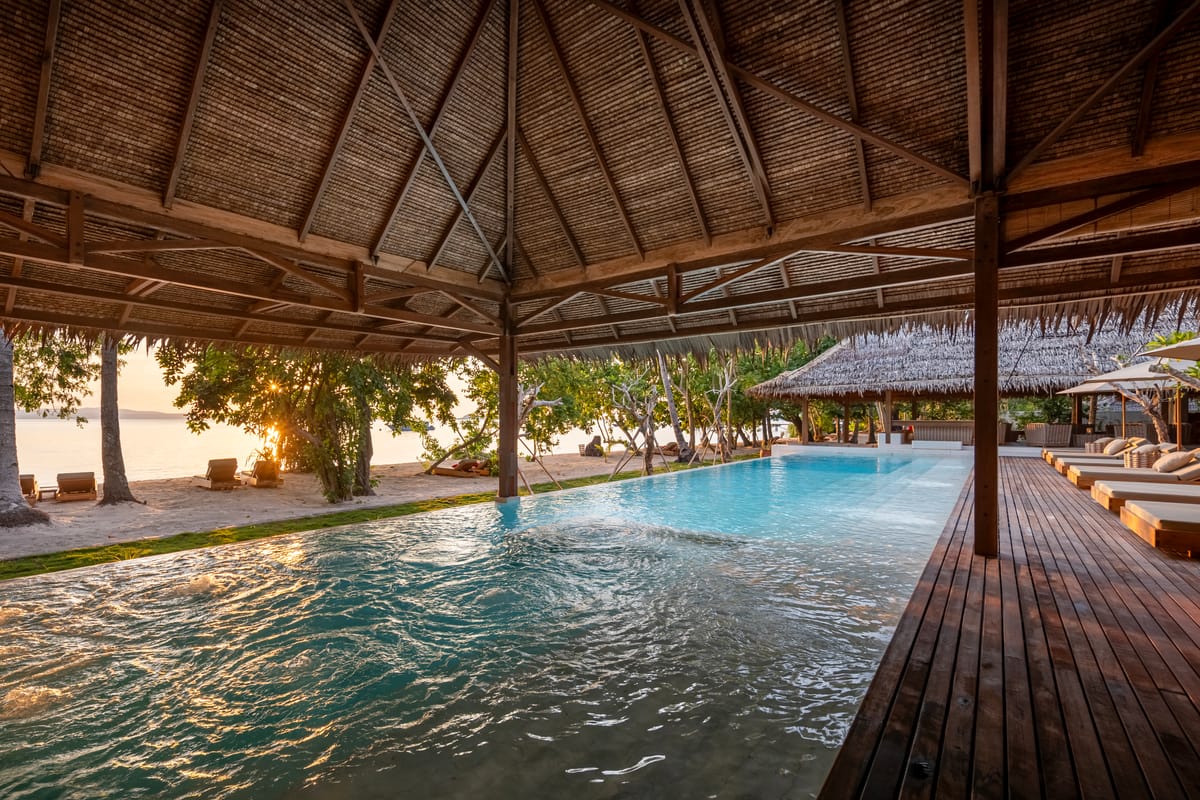 Beachfront infinity pool with a built-in jacuzzi under a grand timber-and-thatch pavilion, sun loungers on a deck overlooking the sea at golden hour on a private island resort in Indonesia