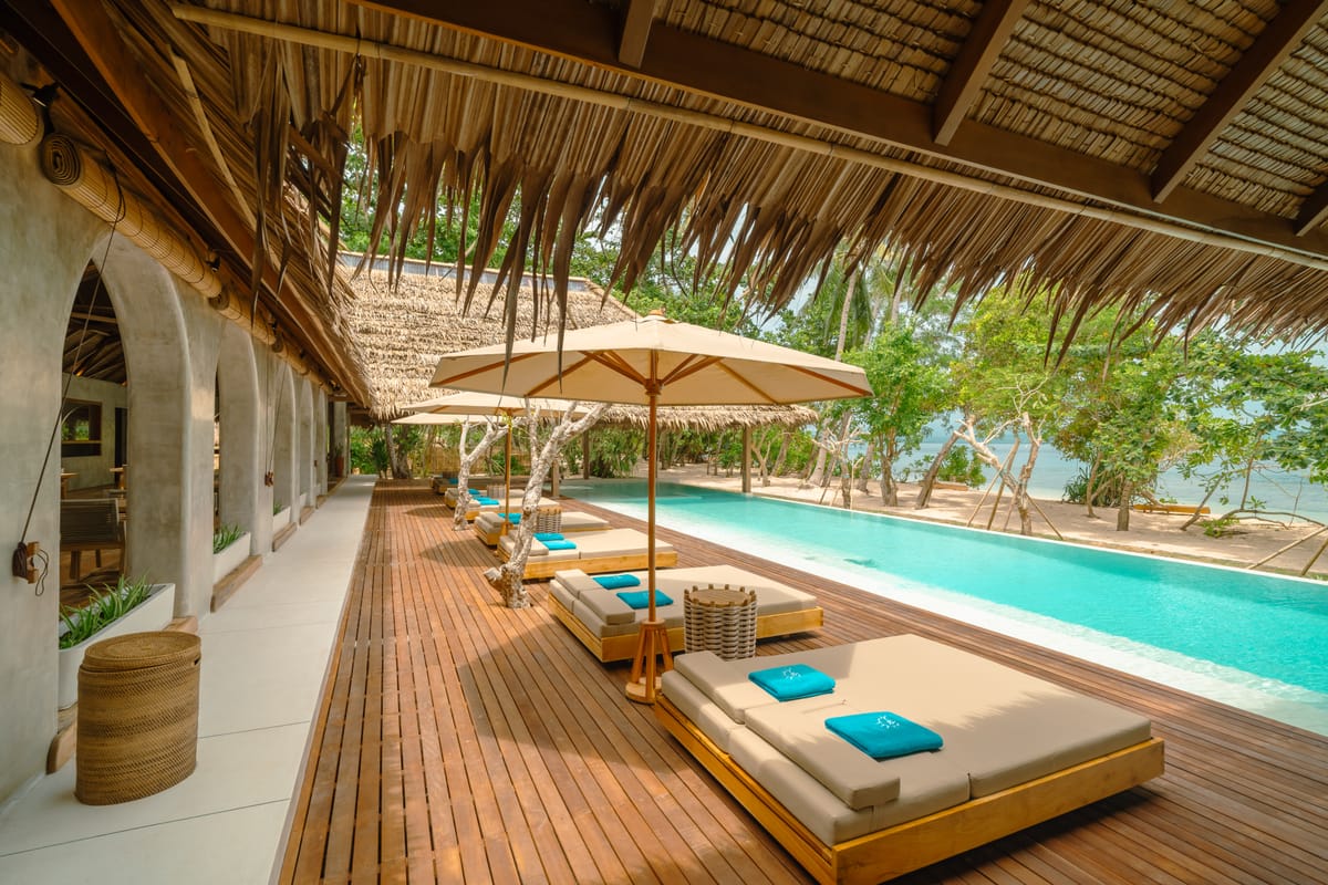 Beachfront infinity pool running alongside a long thatched pavilion with timber daybeds and parasols, opening onto the beach at a private island resort in Indonesia