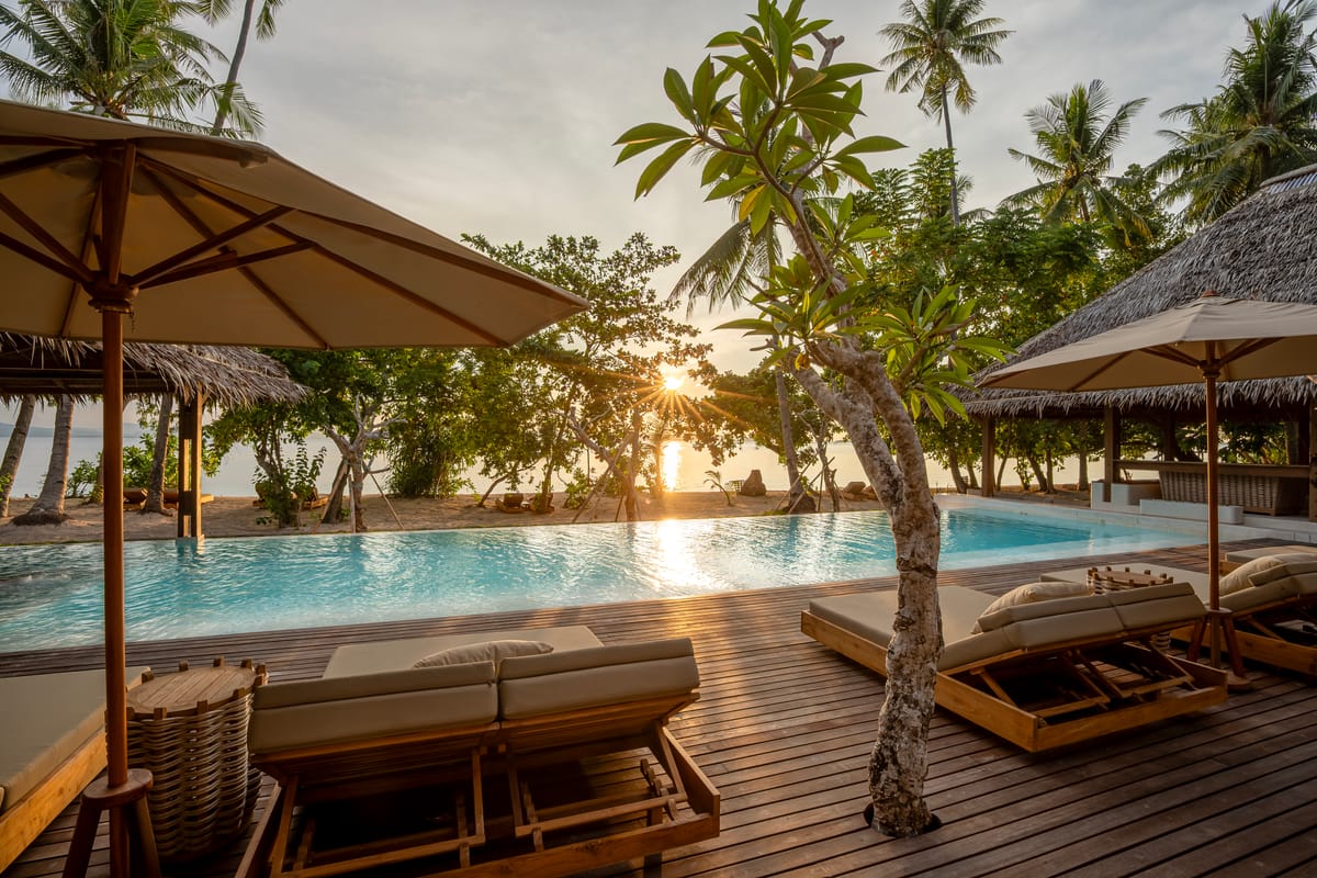 Beachfront resort pool at sunset with timber daybeds, parasols and palms, the sun setting over the sea at a private island resort in Indonesia