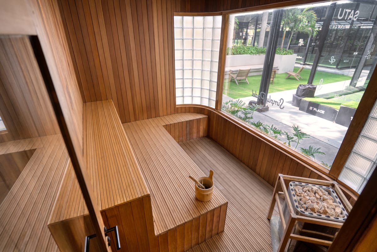 Timber-clad sauna with L-shaped benches and a stone heater beside glass-block and picture windows, built by Tropical Pool at a wellness club in Bali