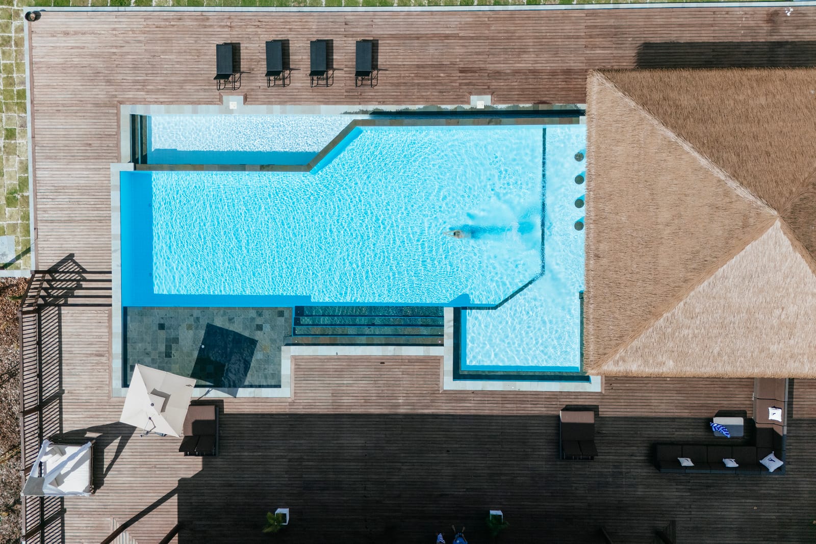 Top-down aerial of a modern geometric pool with a swimmer, timber deck and thatched pavilion in Bali